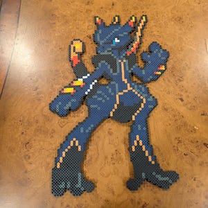 May include: A dark blue pixel art figure with orange and yellow accents. The figure is in a dynamic pose, with arms raised and legs spread. The artwork is made from small, colorful beads.