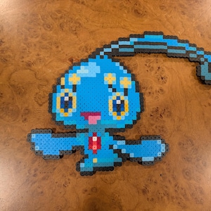 Può includere: Una figura in pixel art realizzata con perline di plastica blu, azzurre, gialle, rosa e rosse. Il personaggio presenta una grande testa arrotondata con una lunga antenna curva, occhi grandi, una piccola bocca rosa e due braccia distese.