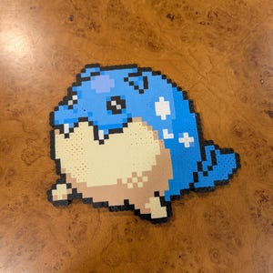 May include: A pixel art creation of a blue and tan creature with black outlines. The artwork features a rounded body with a light blue top, tan belly, and small tail. White cross and dot details are visible.
