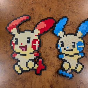 May include: Two pixel art figures crafted from small plastic beads. The left figure is yellow with red ears, red cheek markings, and a red tail. The right figure is yellow with blue ears, blue cheek markings, and a blue tail. Both have black outlines.