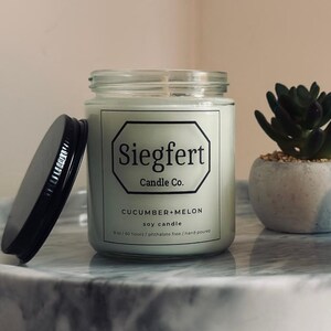 May include: A glass jar candle with a black lid, labeled "Siegfert Candle Co." with "Cucumber + Melon" scent. Two small potted succulents sit beside the candle on a marble tray. The candle is a soy candle.