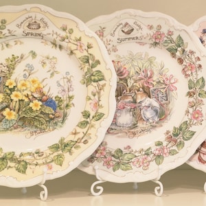 May include: A set of four ceramic plates featuring Brambly Hedge illustrations for Spring, Summer, Autumn, and Winter. Each plate has a scalloped edge, seasonal floral and fruit motifs, and detailed scenes of mice in nature with text labels.