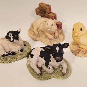 May include: A set of four animal figurines including a black and white lamb on green grass, a pink piglet resting on straw near a brick wall, a black and white calf on green grass, and a yellow chick perched on a textured circular base.