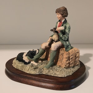 Puede incluir: Una figura pintada a mano que muestra a una persona con chaqueta verde y botas oliva sentada sobre una paca de heno, alimentando a un cordero con biberón. Un border collie blanco y negro descansa a los pies sobre una base de madera oscura.