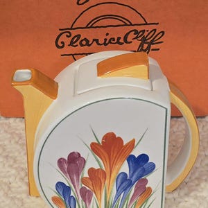 May include: A ceramic teapot with a flat-sided, semi-circular design featuring a hand-painted floral pattern of orange, purple, and blue crocuses. The teapot has a yellow handle, spout, and lid finial. The background text reads Bizarre Clarice Cliff.