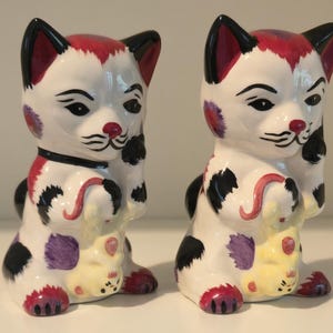 Puede incluir: Dos figuras de gato de cerámica con base blanca y detalles pintados en negro, morado y rojo. Cada gato tiene orejas puntiagudas, un collar negro y sostiene una pequeña criatura amarilla en sus patas delanteras. Tienen rasgos faciales.