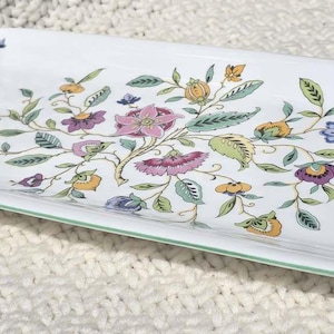 Può includere: Un vassoio da portata rettangolare in ceramica bianca con un colorato motivo floreale composto da fiori e foglie rosa, gialli, blu e verdi. Il vassoio presenta un sottile bordo verde lungo il perimetro e una finitura liscia e lucida.