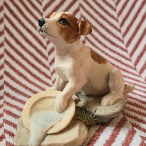 Puede incluir: Una pequeña figura de un cachorro Jack Russell Terrier blanco y marrón sentado sobre una base de roca texturizada. El cachorro tiene una pata apoyada en un cuenco circular de color crema que contiene un líquido azul claro y mira hacia arriba.