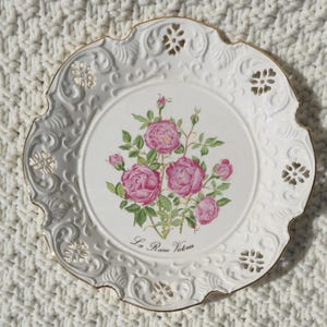 Puede incluir: Un plato decorativo de cerámica blanca con una ilustración central de rosas rosas con hojas verdes y el texto La Reine Victoria. El plato tiene un borde ornamentado en relieve con detalles calados intrincados y un borde dorado.