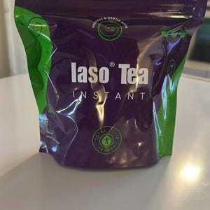 May include: A purple and green stand-up pouch of Iaso Tea Instant. The front features the white text Iaso Tea Instant, the phrase Provides a Gentle Detox, and a circular Total Life Herbal logo. The packaging has a zip-top closure at the top.