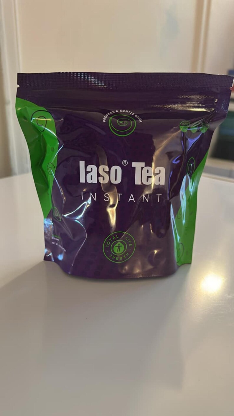 May include: A purple and green stand-up pouch of Iaso Tea Instant. The front features the white text Iaso Tea Instant, the phrase Provides a Gentle Detox, and a circular Total Life Herbal logo. The packaging has a zip-top closure at the top.