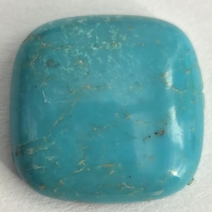 May include: A polished, square-shaped turquoise gemstone. The stone has a smooth surface with natural veining and subtle variations in color, ranging from light to medium blue. The gemstone is a solid, opaque material.