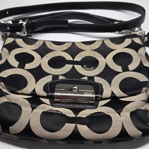 May include: Black and tan Coach shoulder bag with a repeating interlocking C pattern. The bag has a black leather strap and a silver clasp. A small leather tag hangs from the strap.