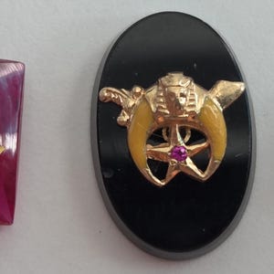 May include: Three vintage Masonic pins. A ruby-colored square pin with a gold Masonic symbol, an oval black pin with a gold and yellow emblem, and a black octagonal pin with a gold emblem.