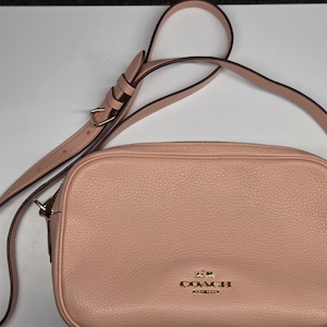 May include: A blush pink Coach crossbody bag with a textured leather exterior. The bag features a long, adjustable strap, gold-tone hardware, and the Coach logo. The bag is rectangular with a zip closure.