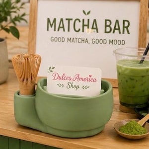 Puede incluir: Un soporte de cerámica verde salvia para batidor de matcha con un batidor de bambú insertado, sosteniendo una tarjeta blanca que dice Dulces America Shop. Al lado hay un vaso de plástico transparente con matcha helado y una pajita negra.
