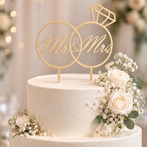 May include: A gold-colored cake topper with the words "Mr & Mrs" in script, set on a white cake decorated with white roses and greenery. The topper features two interlocking rings and a diamond-shaped accent, perfect for a wedding cake.