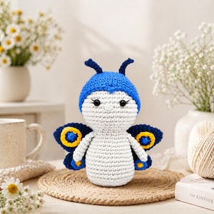 May include: A handmade crochet butterfly doll with a blue cap, white body, and blue and yellow wings. The doll has black eyes and is sitting on a round, woven mat. Decorative flowers and a mug are in the background.