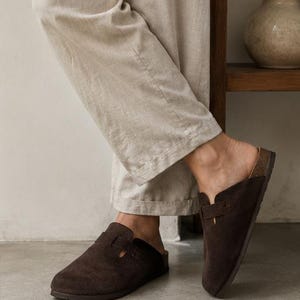 Könnte beinhalten: Ein Paar dunkelbraune Wildleder-Clogs zum Hineinschlüpfen mit abgerundeter Zehenpartie, einem breiten Riemen über dem Spann mit Ausschnitt und einer Zwischensohle in Korkoptik. Das Schuhwerk hat eine flache Sohle und ein offenes Fersendesign.