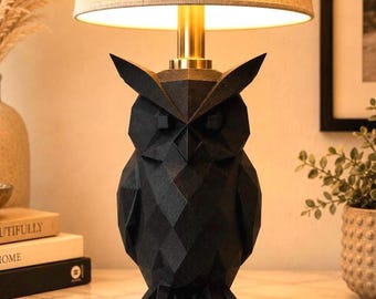 May include: A table lamp featuring a black, geometric, faceted owl-shaped base. The lamp has a cylindrical, light-coloured fabric lampshade and a gold-toned metal neck connecting the owl base to the light source, emitting a warm glow.