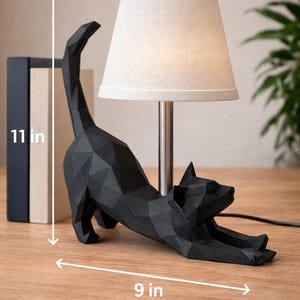 K&ouml;nnte beinhalten: Schwarze Katzen-Tischlampe mit beigem Lampenschirm. Die Katze befindet sich in einer Dehnposition und misst 23 cm lang und 28 cm hoch. Die Lampe hat einen silbernen Sockel und ein schwarzes Netzkabel. Zwei B&uuml;cher sind im Hintergrund zu sehen.