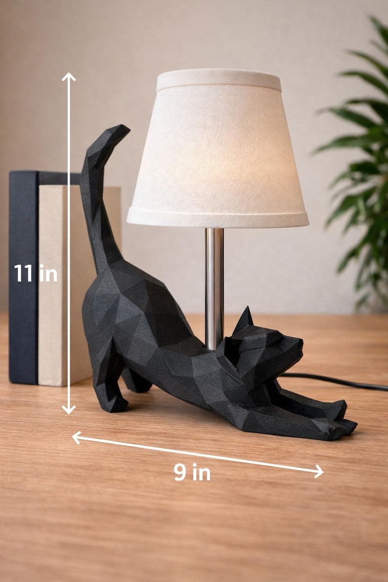 K&ouml;nnte beinhalten: Schwarze Katzen-Tischlampe mit beigem Lampenschirm. Die Katze befindet sich in einer Dehnposition und misst 23 cm lang und 28 cm hoch. Die Lampe hat einen silbernen Sockel und ein schwarzes Netzkabel. Zwei B&uuml;cher sind im Hintergrund zu sehen.