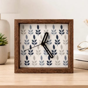 May include: Square wooden clock with a white face featuring a pattern of blue and gray leaves. The clock has black hands and a dark brown wooden frame. The clock is approximately 6 inches tall and wide.