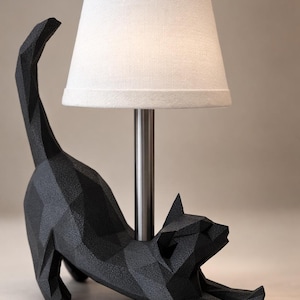 K&ouml;nnte beinhalten: Schwarze, geometrische Katzen-Tischlampe mit wei&szlig;em Lampenschirm. Die Katze befindet sich in einer Dehnposition, mit offenem Maul. Die Lampe hat einen silbernen Metallpfosten und steht auf einer hellen Oberfl&auml;che. Ca. 30 cm hoch.