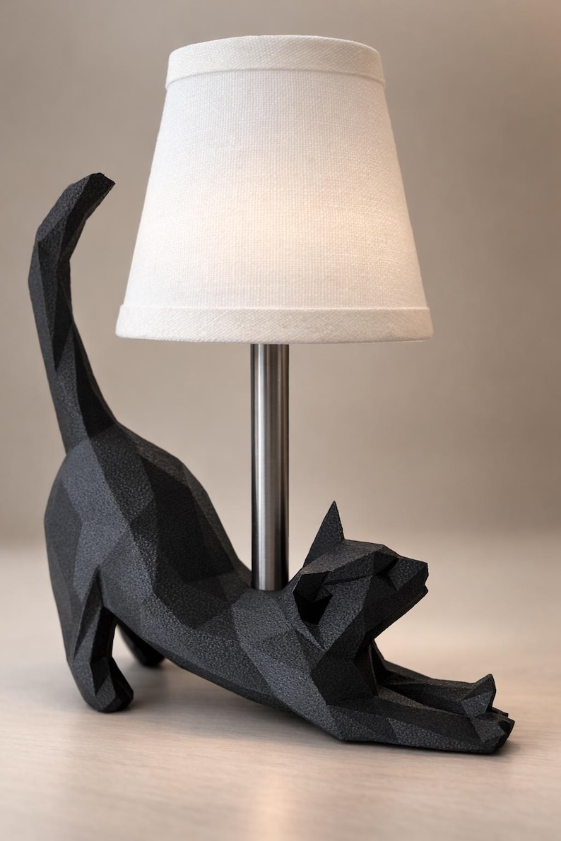 K&ouml;nnte beinhalten: Schwarze, geometrische Katzen-Tischlampe mit wei&szlig;em Lampenschirm. Die Katze befindet sich in einer Dehnposition, mit offenem Maul. Die Lampe hat einen silbernen Metallpfosten und steht auf einer hellen Oberfl&auml;che. Ca. 30 cm hoch.