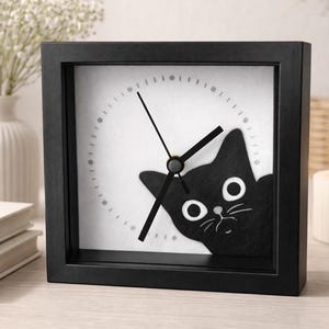 May include: A square black clock with a white face featuring a black cat peeking from the side. The clock has black hands and a circular time marker. The clock is set on a light-colored surface.