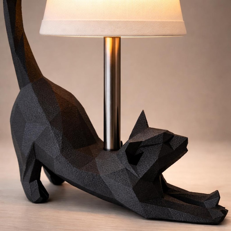K&ouml;nnte beinhalten: Eine schwarze, geometrische Katzen-Tischlampe mit cremefarbenem Lampenschirm. Die Katze befindet sich in einer Dehnposition, mit ausgestreckten Vorderpfoten. Die Lampe hat eine silberne Metallstange.