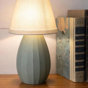 May include: A table lamp with a matte sage green, vertically ribbed, egg-shaped base and a tapered, off-white fabric lampshade. The lamp emits a warm, soft glow, illuminating the textured surface of the base and the surrounding area.