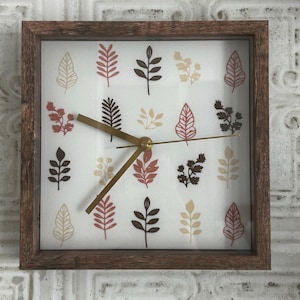 May include: Square wall clock with a brown wooden frame. The clock face is white with a pattern of colorful leaves in shades of red, brown, and beige. The clock hands are gold.