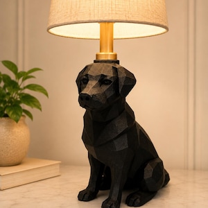 May include: A table lamp featuring a black, geometric, faceted dog-shaped base. The lamp has a cylindrical brass-colored neck and a beige, textured fabric drum lampshade that emits a warm, soft glow from the light source inside the shade.