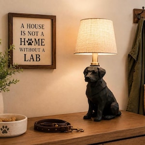 May include: A table lamp with a black geometric Labrador dog base and a beige fabric lampshade. Next to it is a framed sign that reads A HOUSE IS NOT A HOME WITHOUT A LAB, a white bowl with a paw print, and a brown leather dog leash.