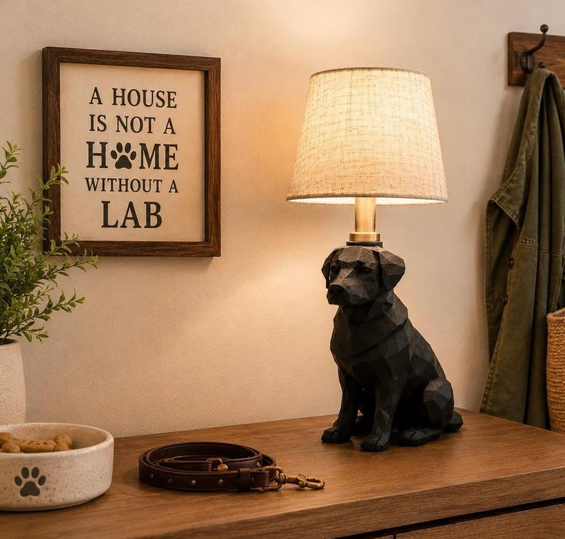 May include: A table lamp with a black geometric Labrador dog base and a beige fabric lampshade. Next to it is a framed sign that reads A HOUSE IS NOT A HOME WITHOUT A LAB, a white bowl with a paw print, and a brown leather dog leash.