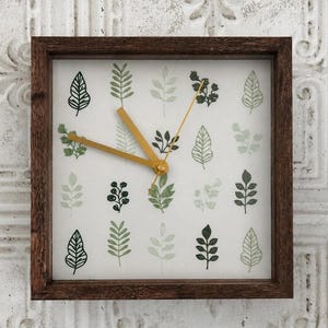 May include: Square wooden wall clock with a dark brown frame. The clock face features a white background with various green leaf illustrations. The clock hands are gold. A decorative piece for a living room or bedroom.