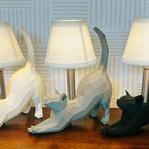 K&ouml;nnte beinhalten: Drei katzenf&ouml;rmige Tischlampen in Wei&szlig;, Grau und Schwarz. Jede Lampe hat einen wei&szlig;en Lampenschirm und einen Metallstiel. Die Katzenfiguren befinden sich in einer Dehnpose, wobei ihre Schw&auml;nze nach oben gebogen sind. Die Lampen stehen auf einer Holzoberfl&auml;che.