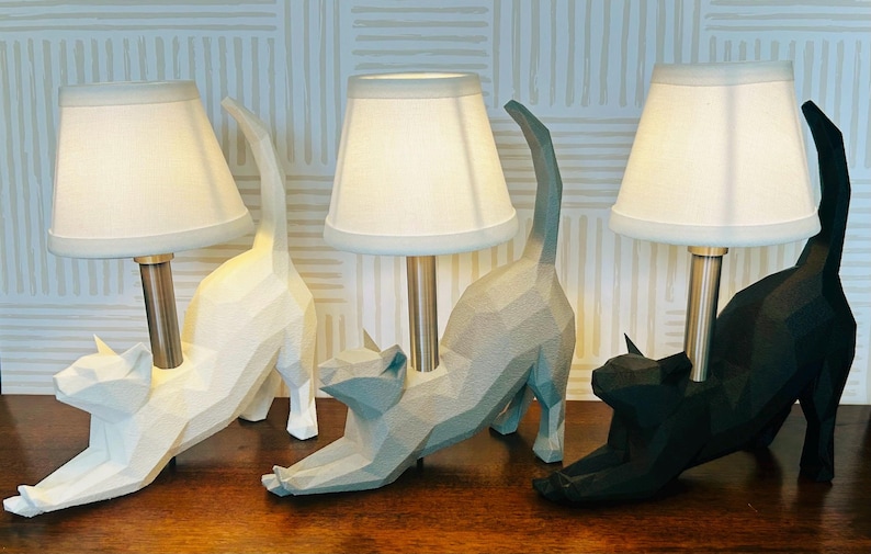 K&ouml;nnte beinhalten: Drei katzenf&ouml;rmige Tischlampen in Wei&szlig;, Grau und Schwarz. Jede Lampe hat einen wei&szlig;en Lampenschirm und einen Metallstiel. Die Katzenfiguren befinden sich in einer Dehnpose, wobei ihre Schw&auml;nze nach oben gebogen sind. Die Lampen stehen auf einer Holzoberfl&auml;che.
