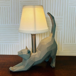 K&ouml;nnte beinhalten: Eine graue, geometrische Katzen-Tischlampe mit cremefarbenem Lampenschirm. Die Katze befindet sich in einer Dehnpose, mit gew&ouml;lbtem Schwanz. Die Lampe hat einen Metallpfosten und steht auf einer dunklen Holzoberfl&auml;che.