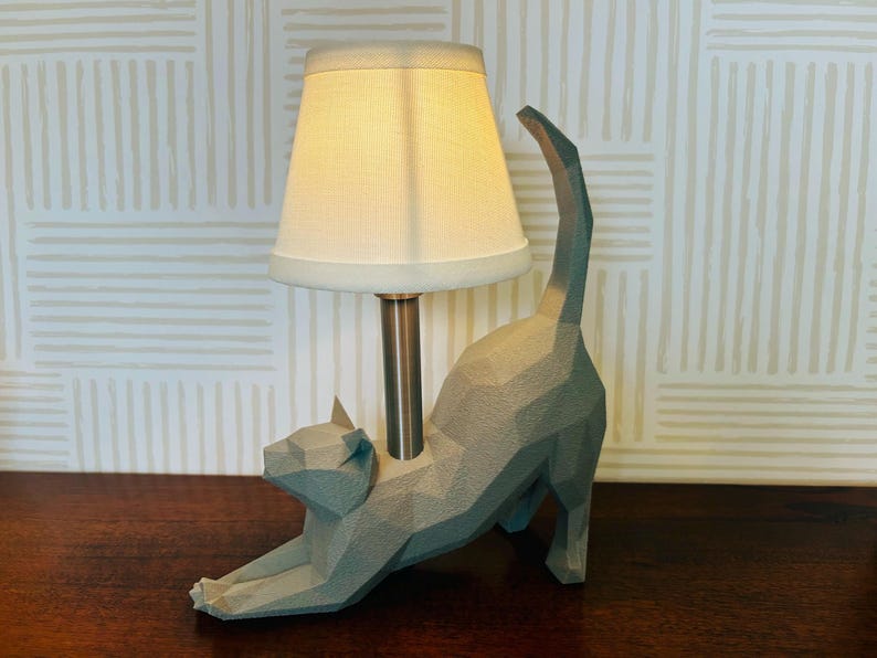 K&ouml;nnte beinhalten: Eine graue, geometrische Katzen-Tischlampe mit cremefarbenem Lampenschirm. Die Katze befindet sich in einer Dehnpose, mit gew&ouml;lbtem Schwanz. Die Lampe hat einen Metallpfosten und steht auf einer dunklen Holzoberfl&auml;che.
