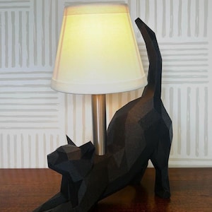 K&ouml;nnte beinhalten: Eine schwarze, geometrische Katzenfigur, die sich dehnt, mit einem cremefarbenen Lampenschirm und einem silbernen Lampenfu&szlig;. Die Katze steht auf einer Holzoberfl&auml;che, mit einem gemusterten Tapetenhintergrund.