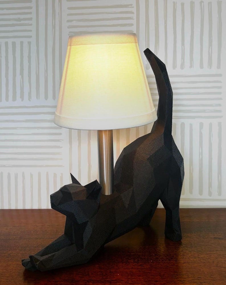K&ouml;nnte beinhalten: Eine schwarze, geometrische Katzenfigur, die sich dehnt, mit einem cremefarbenen Lampenschirm und einem silbernen Lampenfu&szlig;. Die Katze steht auf einer Holzoberfl&auml;che, mit einem gemusterten Tapetenhintergrund.