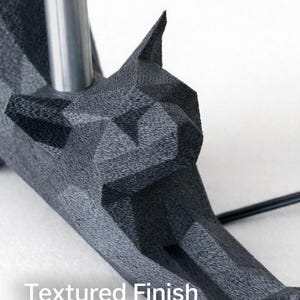 K&ouml;nnte beinhalten: Nahaufnahme eines dunkelgrauen, geometrisch geformten Objekts mit strukturierter Oberfl&auml;che. Das Objekt hat ein wolfs&auml;hnliches Kopfdesign und eine silberne Metallstange. Der Text "Textured Finish" ist sichtbar.