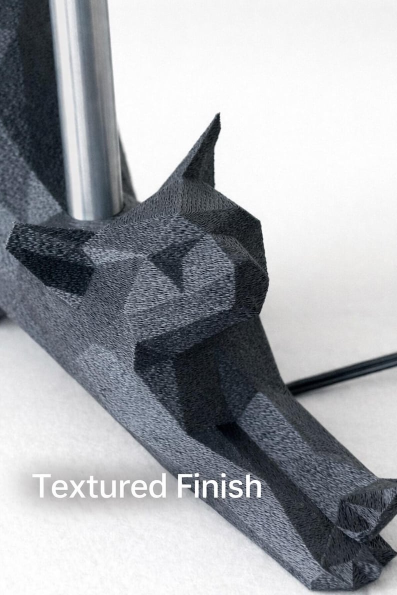 K&ouml;nnte beinhalten: Nahaufnahme eines dunkelgrauen, geometrisch geformten Objekts mit strukturierter Oberfl&auml;che. Das Objekt hat ein wolfs&auml;hnliches Kopfdesign und eine silberne Metallstange. Der Text "Textured Finish" ist sichtbar.