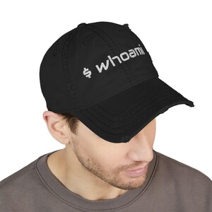 May include: A black distressed baseball cap featuring the white embroidered text "$ whoami" on the front panel. The hat has a curved brim with frayed edges and a structured crown with visible stitching details across the fabric panels.
