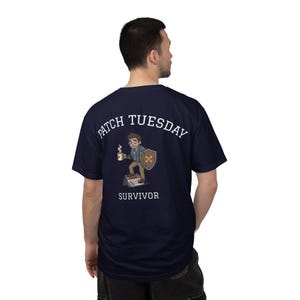 May include: A navy blue short-sleeve t-shirt featuring a graphic on the back. The design shows a cartoon character holding a steaming mug and a shield with bandages, standing on a calendar. The text reads PATCH TUESDAY SURVIVOR in white.