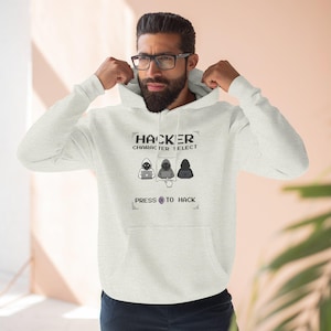 May include: A light grey hoodie featuring the text "HACKER CHARACTER SELECT" above three pixelated hacker figures. Below the figures is the text "PRESS TO HACK". The hoodie has a drawstring hood and a front pocket.