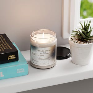Könnte beinhalten: Eine brennende Legacy System Kerze in einem durchsichtigen Glas mit schwarzem Deckel, neben einer kleinen Topf-Sukkulente und zwei Büchern. Das Kerzenetikett enthält Text. Die Kerze steht auf einem weißen Regal.