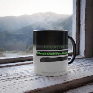 May include: A black and white color-changing mug with the text "Top Secret: I Break Stuff For A Living" in green. The mug's design includes a black band and the phrase "Redacted Until Warm". Steam rises from the mug.